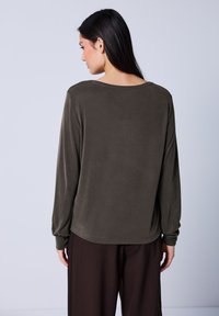 Langärmliges Shirt in dunklem Olivgrün, aus weichem Stoff gefertigt, mit einem weiten Ausschnitt und schmal zulaufenden Ärmeln. Kombiniert mit gestreiften dunklen Hosen.