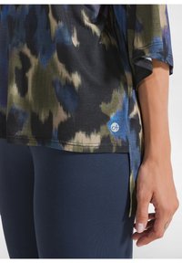 Top a maniche corte in stampa astratta nera, blu e verde; include fessure laterali e un dettaglio del logo blu vicino all'orlo.
