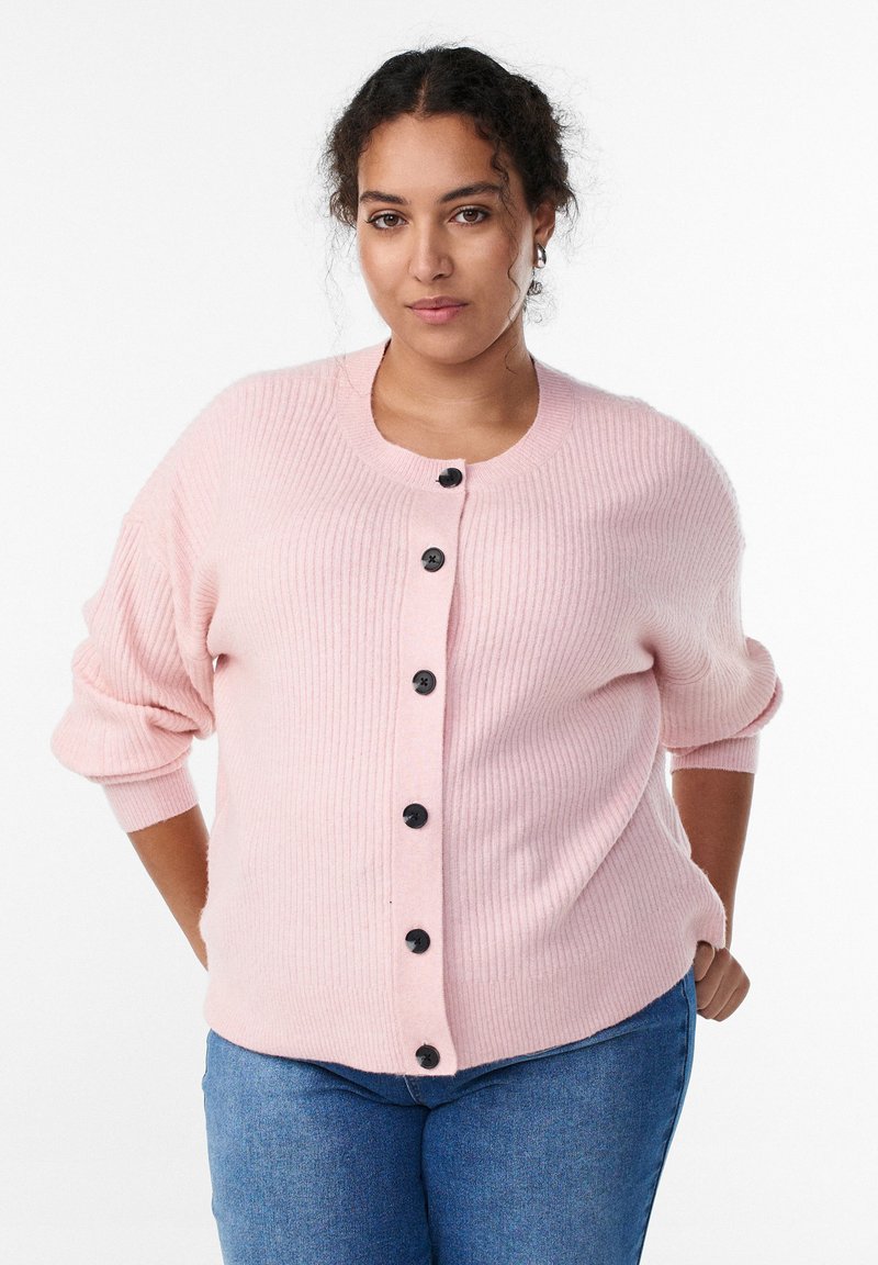 Gaiši rozā ribota kardigans ar apaļu izgriezumu, melnām pogām un nedaudz uzpūstām piedurknēm, kas valkāts virs zilām džinsu biksēm.