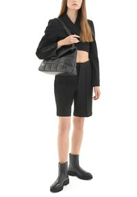 Giacca nera a lunghezza crop con scollatura profonda, pantaloni neri su misura e stivali alla caviglia neri. La borsa è trapuntata con una tracolla a catena.