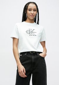 Biele bavlnené tričko s čiernym logom Calvin Klein, doplnené o čierne rifle s vysokým pásom a čierny opasok so zlatou prackou.