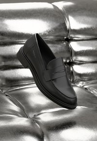Black leather loafer featuring a polished finish, rounded toe, and a decorative strap across the vamp, set against a metallic tufted background.