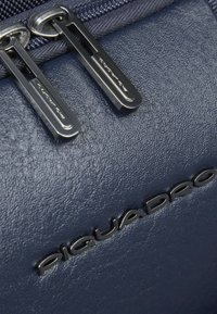 Marineblaue Ledertasche mit strukturierter Oberfläche, ausgestattet mit silbernen Reißverschlüssen und geprägtem "Piquadro"-Logo. Nahaufnahme von Beschlägen und Materialdetails.