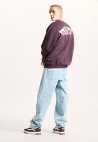 Fioletowy sweter z okrągłym dekoltem z logo Vans z tyłu, w połączeniu z jasnoniebieskimi luźnymi dżinsami i brązowymi sneakersami z białymi akcentami.