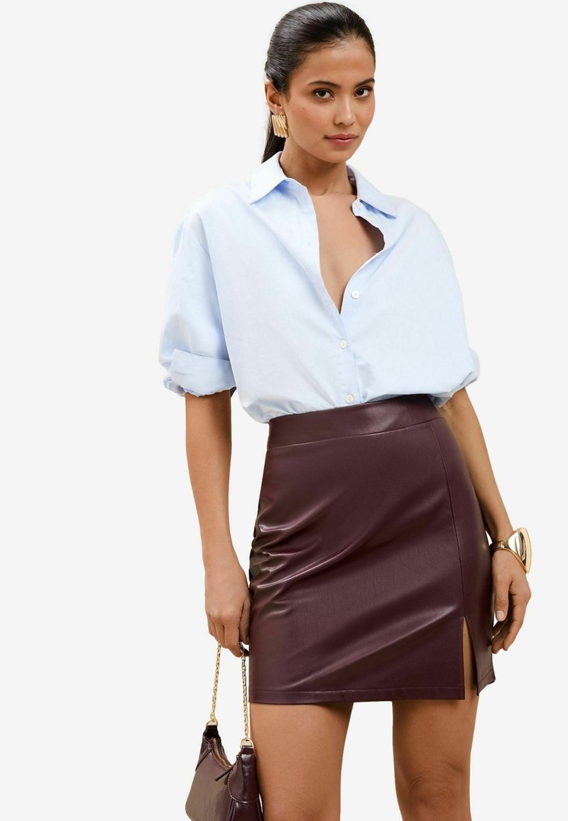 Lichtblauw oversized button-up shirt met opgerolde mouwen, gecombineerd met een strakke bordeauxrood neplederen mini-rok met een zijsplit. Gouden accessoires.