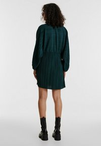 Grünes plissiertes Kleid mit elastischem Taillenbund, langen Ärmeln und lässiger Passform, kombiniert mit schwarzen klobigen Knöchelstiefeln.