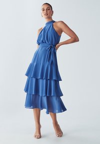 Plava halter haljina s višeslojnim dizajnom, koja se sastoji od preklapajućih slojeva i kravate oko struka. Izrađena od laganog, lepršavog materijala.
