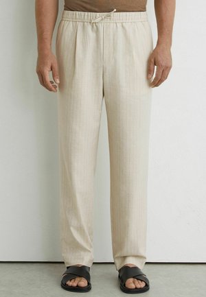 Mężczyzna w beżowych spodniach ze ściągaczem z subtelnymi paskami i czarnych sandałach z odkrytymi palcami, stojący przed jasną ścianą.