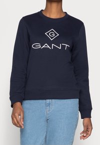 Osoba ubrana w granatowy polar z białym logo "GANT" i jasnoniebieskie dżinsy, stoi przed jednolitym tłem.