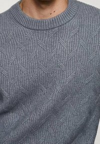 Szary sweter dziany z teksturowanym wzorem nakładających się na siebie liter V, zaokrąglony dekolt i ściągacze przy rękawach. Wykonany z miękkiego, grubego materiału.