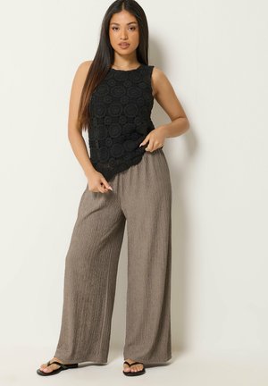 Femme aux longs cheveux portant un top en crochet noir sans manches, un pantalon large ample taupe texturé et des sandales plates noires, debout contre un mur blanc.