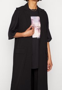 Dlouhá černá vesta z hladké látky, s límcem, předními kapsami a otevřeným střihem. Nošena přes černé tričko s grafickým potiskem.