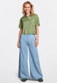 Grünes, blumiges kurzärmliges Hemd kombiniert mit blauen, weit geschnittenen Jeans mit Knopfdetails an den Seiten, getragen mit grünen und gelben Sneakers.
