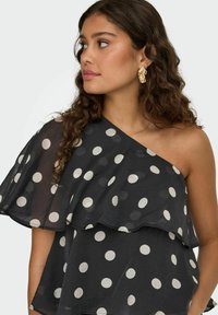 Femme aux longs cheveux bouclés portant de grandes boucles d'oreilles en or et une robe noire asymétrique à pois blancs, regardant sur le côté.