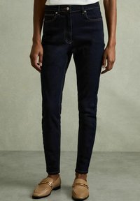 Mörkblå jeans i slim fit med orange sömmar, fem fickor och knappgylf, kombinerat med beige loafers.