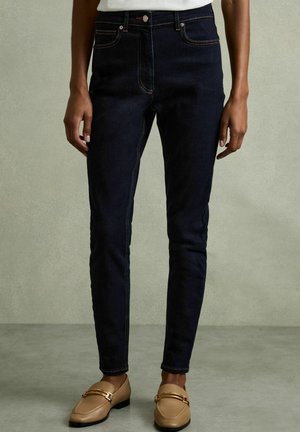 Jeans in denim blu scuro a taglio slim, con cuciture arancioni, cinque tasche e chiusura con bottone, abbinati a mocassini beige.
