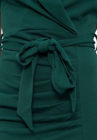 Robe en tissu vert foncé avec un nœud noué à la taille et des plis froncés mettant en valeur la silhouette ajustée au niveau de la taille.
