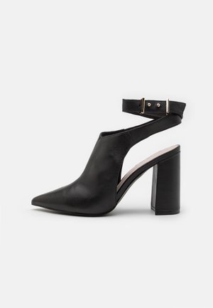 Black leather heeled shoe with pointed toe and open side design. Features an ankle strap secured with a gold buckle. Block heel design.