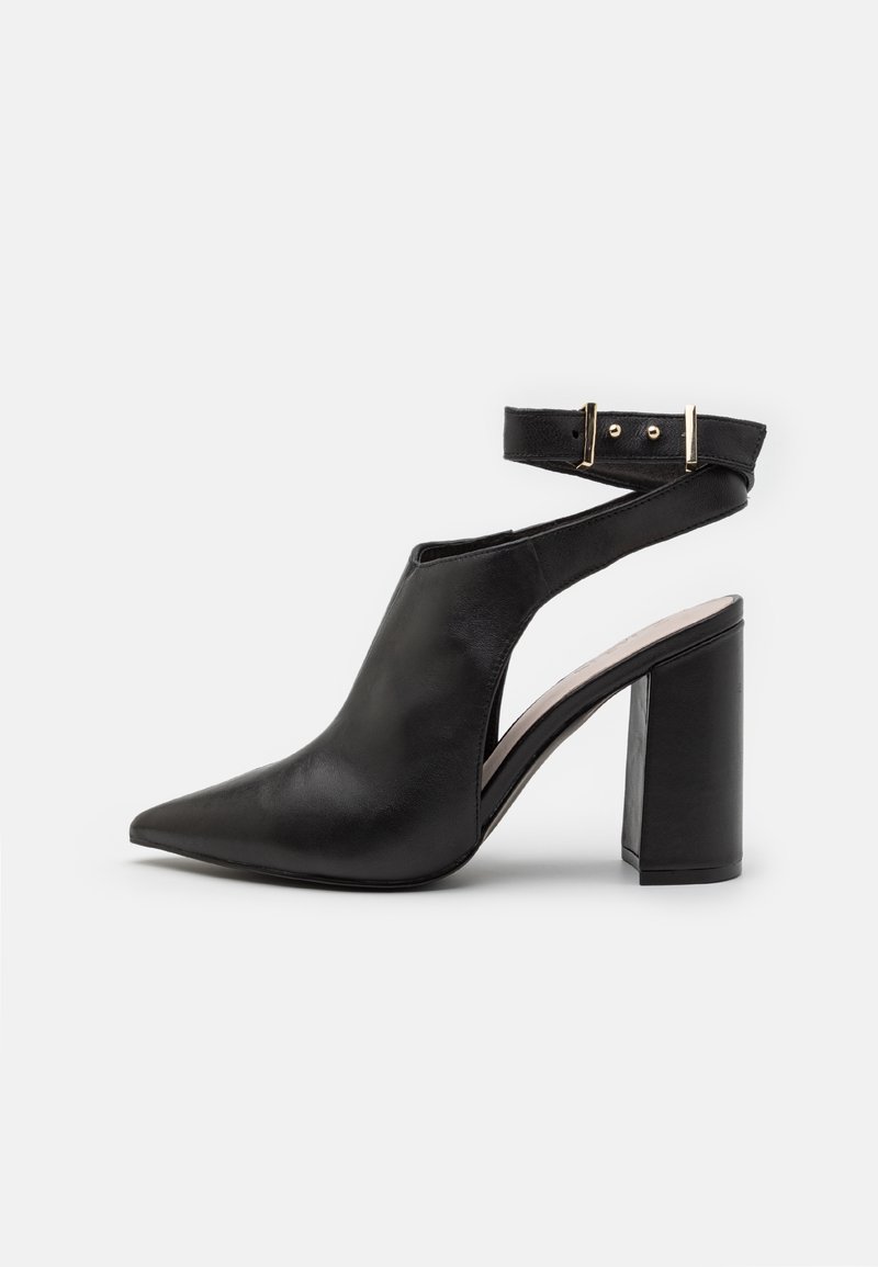 Chaussure à talon en cuir noir avec bout pointu et design latéral ouvert. Présente une bride à la cheville fixée par une boucle dorée. Talon bloc.