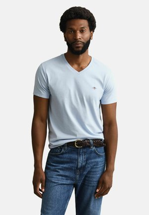 Man met baard en afro-kapsel, gekleed in een lichtblauw V-hals T-shirt en blauwe spijkerbroek met een gevlochten bruine riem.