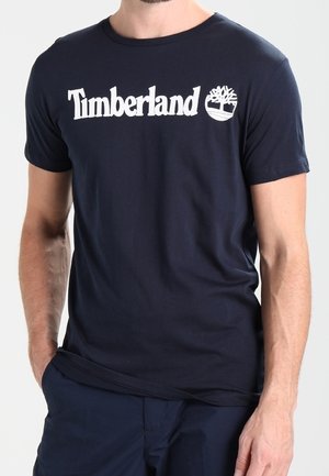 Man wearing navy blue Timberland t-shirt with white logo and tree emblem, with one hand in pocket and neutral background.