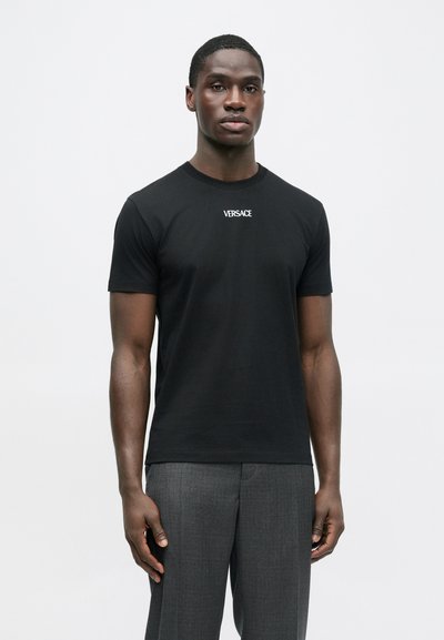 Μαύρο βαμβακερό μπλουζάκι με στρογγυλή λαιμόκοψη και κοντά μανίκια, με λευκή επιγραφή "Versace" στο στήθος. Φορεμένο με γκρι παντελόνι ραμμένο στα μέτρα.