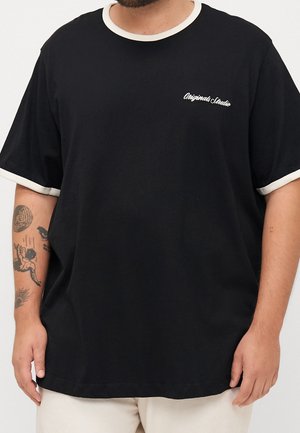 Homme portant un T-shirt noir avec un liseré blanc sur le col et les manches, avec le texte "Originals Studio" sur la poitrine, montrant des tatouages sur l'avant-bras gauche.