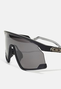 Schwarze Sportsonnenbrille mit großen grauen Gläsern und einem geometrischen Rahmen. Verfügt über flexible Gummiakzente und ein metallisches Logo-Detail an der Seite.