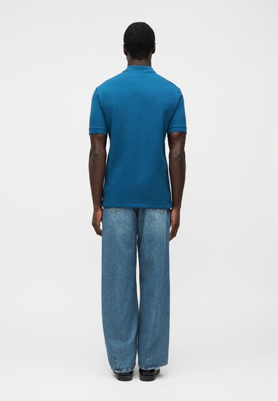 Τυρκουάζ πόλο μπλούζα με κοντά μανίκια και ριμπ γιακά, σε συνδυασμό με ανοιχτόχρωμο τζιν παντελόνι φαρδιάς γραμμής. Λείο ύφασμα, άνετη εφαρμογή.