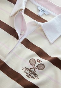Polo beige et marron à rayures avec col, boutons et raquettes de tennis croisées brodées, couronne et texte "Tennis & Paddle The Grand Pasadena Club."