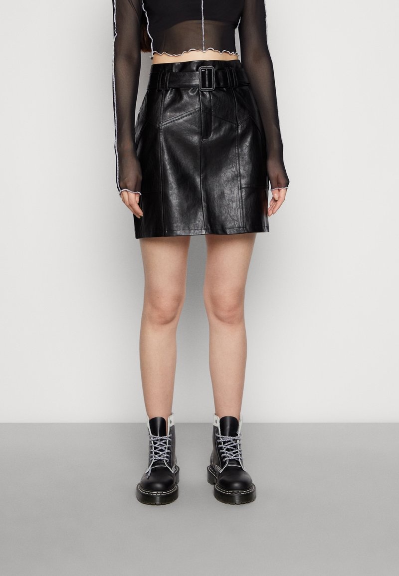 Personne portant une jupe en cuir noire ceinturée, un top court transparent à manches longues et des bottes de combat noires à lacets, debout sur un sol gris.