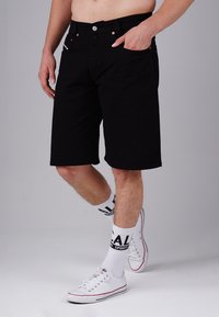 Black denim shorts with a straight cut, five-pocket design, copper rivets, and a button closure. Paired with white socks and sneakers.
