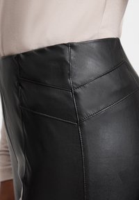 Jupe en simili cuir noir avec une taille haute, un léger éclat et des coutures détaillées, dotée de deux poches arrière inclinées.