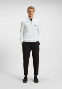 Weißer Reißverschluss-Pullover mit strukturiertem Vorderpanel und schwarzem "BOSS"-Logo, kombiniert mit schwarzen, maßgeschneiderten Hosen und schwarzen Sportschuhen.