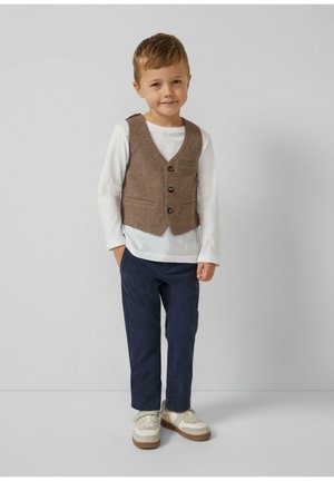 Jeune garçon debout les mains dans les poches, portant un gilet marron à boutons, une chemise blanche à manches longues, un pantalon bleu marine et des baskets blanches.