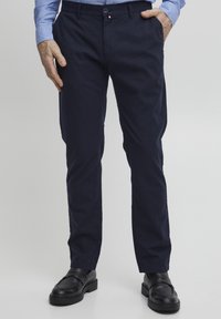 Pantaloni in cotone blu navy con vestibilità slim, dotati di tasche laterali e un piccolo patch con logo. Abbinati a scarpe in pelle nere per un contrasto.