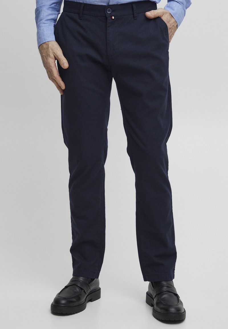 Pantaloni in cotone blu navy con vestibilità slim, dotati di tasche laterali e un piccolo patch con logo. Abbinati a scarpe in pelle nere per un contrasto.