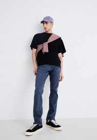 Zwart t-shirt, lichtpaarse baseballpet, roze trui om de schouders gebonden, blauwe jeans, zwarte sneakers met witte veters, casual outfit.