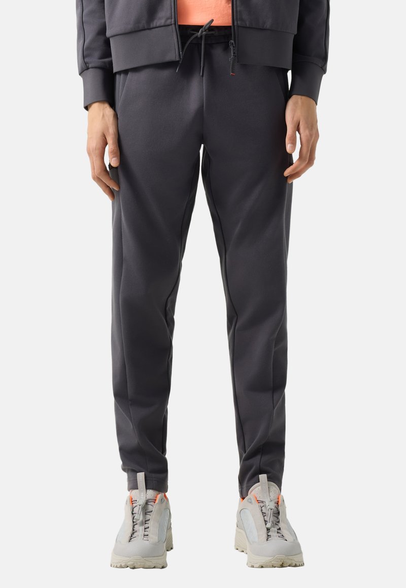 Osoba ubrana w ciemnoszare spodnie dresowe z sznurkiem w pasie oraz jasnoszare buty sportowe z pomarańczowymi akcentami.