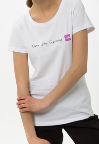 Bílé tričko s krátkým rukávem se ručně psaným textem "Nikdy nepřestávej objevovat" a malým fialovým logem na levé straně.