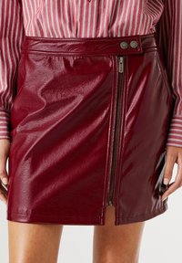 Jupe mini en simili cuir bordeaux avec fermeture éclair sur le côté, texture lisse et finition brillante contrastante. Accentuée par des boutons-pression.