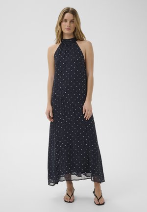 Femme portant une robe longue noire sans manches à pois blancs et des sandales noires, debout sur un fond blanc uni.