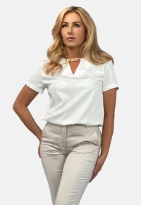 Biała bluzka z krótkim rękawem, z dekoltem w kształcie V i złotymi akcentami. Połączona z beżowymi dopasowanymi spodniami, które podkreślają gładki materiał i czyste linie.