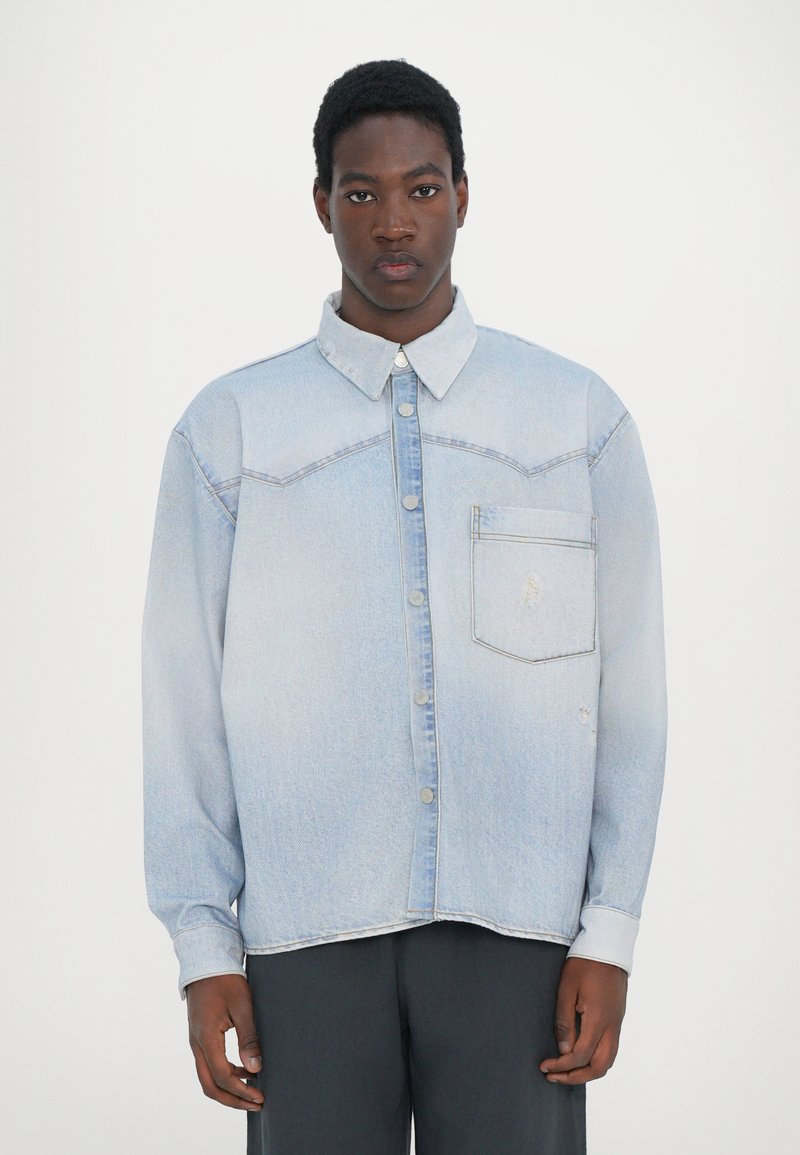 Lyseblå denim overhemse med knappestolpe foran, brystlomme og spiss krage. Har falmet tekstur og lett slitasje langs kantene.