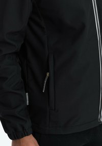 Veste noire avec une fermeture éclair sur le devant, des poignets élastiques et une poche latérale avec une fermeture éclair. Le tissu a une finition texturée.