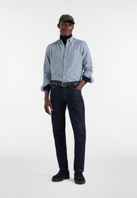 Uomo che indossa un berretto verde, una camicia a quadri blu con bottoni sopra un dolcevita blu scuro, jeans scuri, una cintura nera e scarpe nere, in piedi con una mano in tasca.