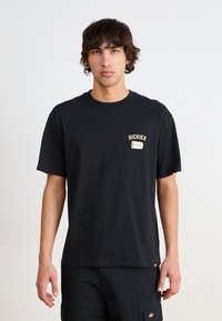 T-shirt en coton noir avec un petit logo graphique beige sur la partie gauche de la poitrine. Manches courtes et design classique à col rond.