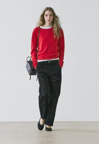 Pull rouge en maille douce, porté sur une chemise blanche. Pantalon noir à jambes larges, associé à des chaussures plates noires. Petit sac à main texturé avec bandoulière.
