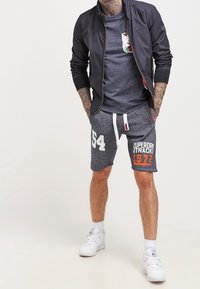 Marineblå bomberjacka, grå t-shirt med blommig ficka, grå shorts med vit "54" och orange "Superdry Track 1972," matchat med vita sneakers.