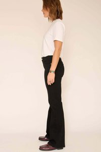 Jean noir évasé taille haute, associé à un t-shirt blanc. Le mannequin est de profil, portant des mocassins bordeaux et des accessoires.